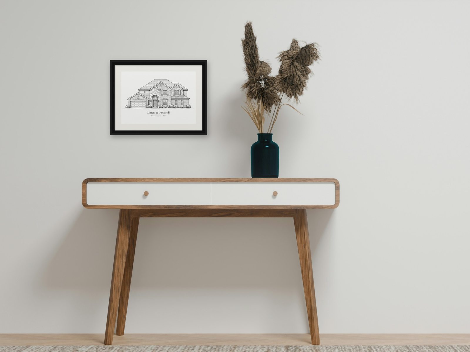 A framed pencil drawing of a two-story brick and stone house, captioned Marcus and Dana Hill, hanging above a wooden console table